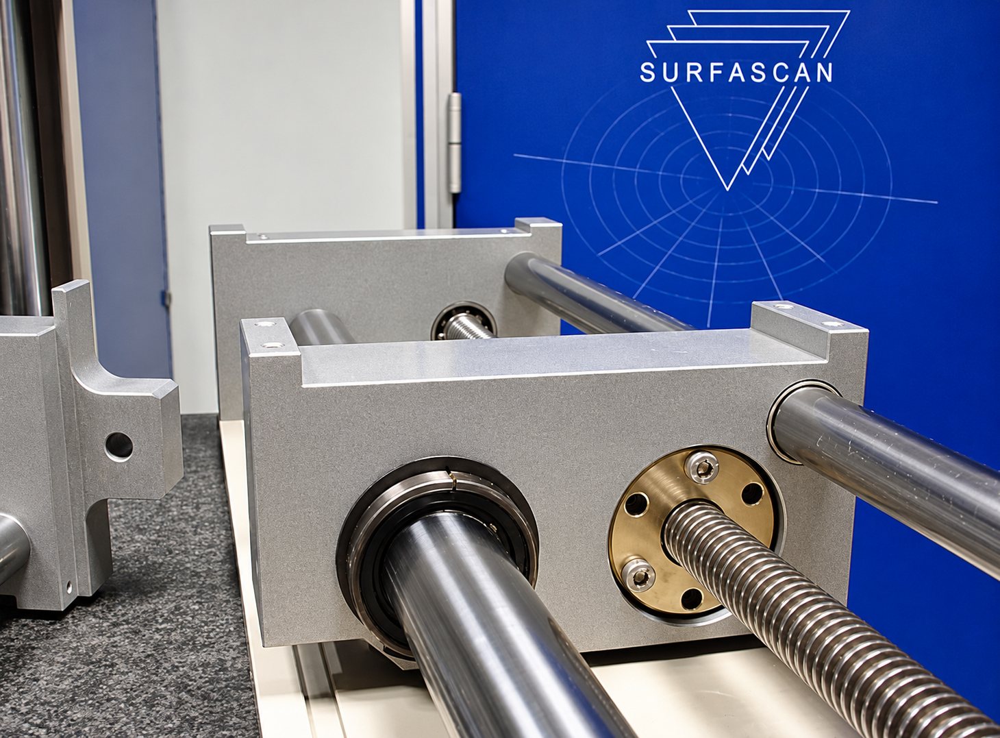 Gros plan sur des blocs de guidage en aluminium montés sur des tiges d'acier polies et une vis mère, devant un panneau bleu portant le logo Surfascan.