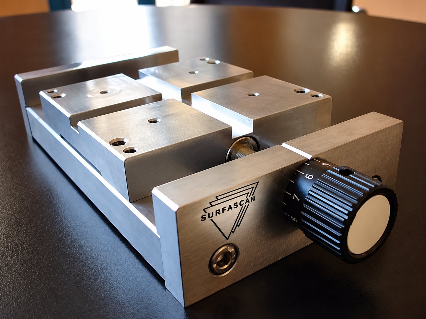 Un étau de précision en aluminium brossé portant la marque Surfascan, avec une molette noire graduée, posé sur une table sombre et brillante.