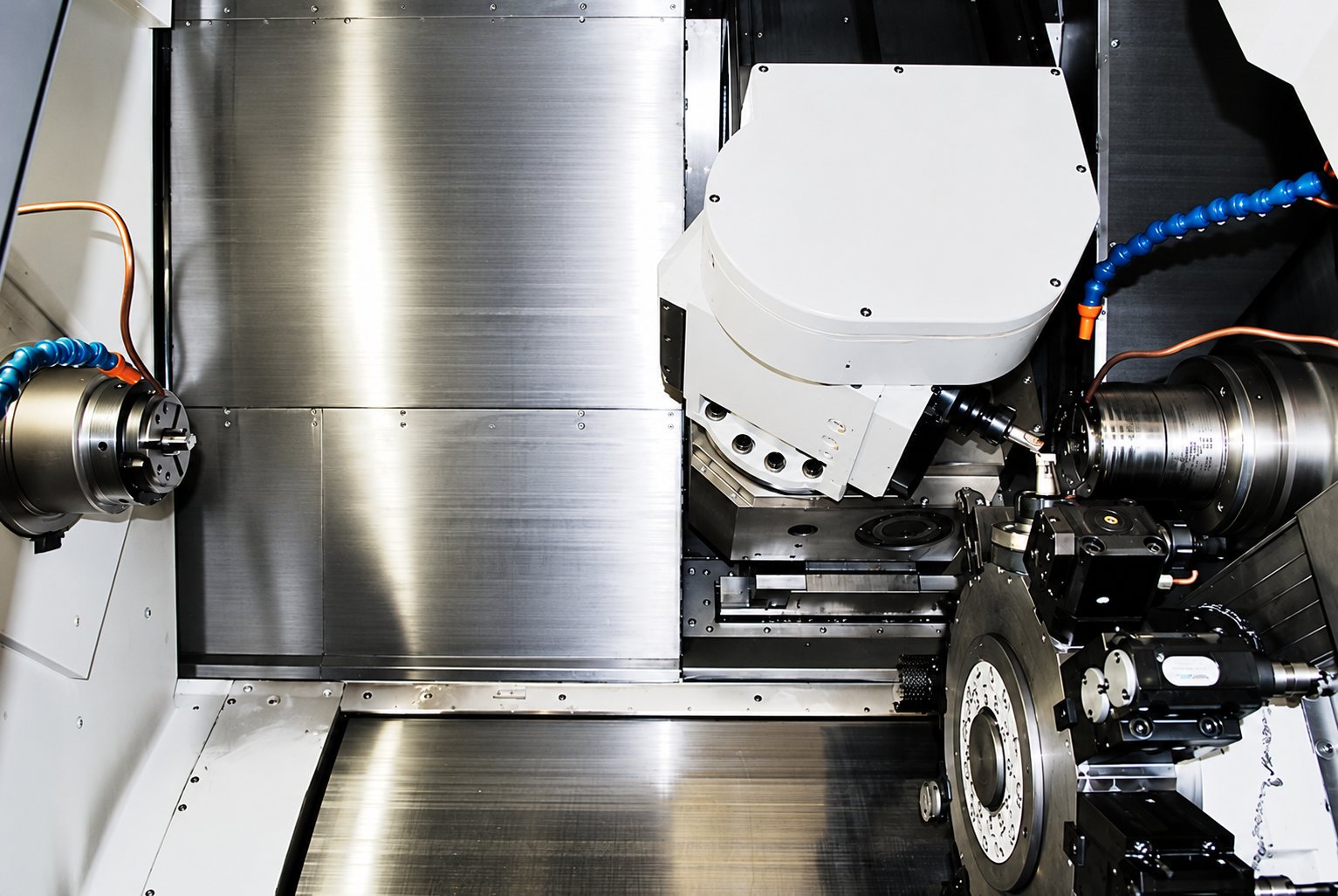 Vue de l'intérieur d'une machine-outil CNC avec parois en acier brillant, une tête d'usinage blanche, des mandrins et des tuyaux de refroidissement bleus.