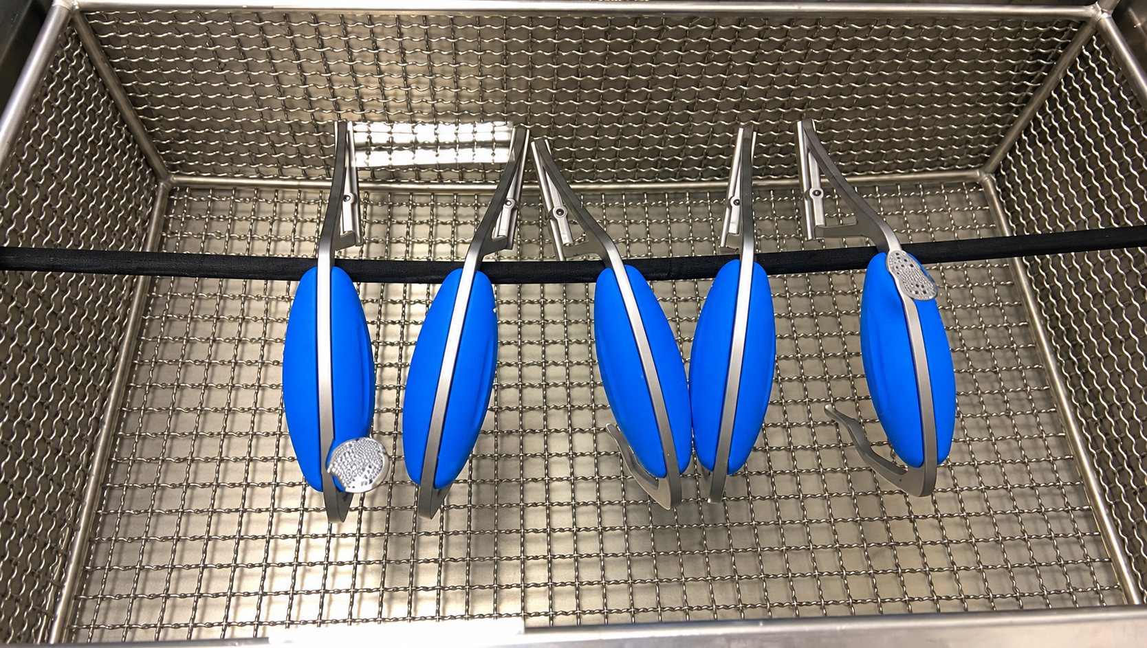 Cinq objets ovales bleus munis de pinces noires sont suspendus à une barre au-dessus d'un bac en métal à fond quadrillé.