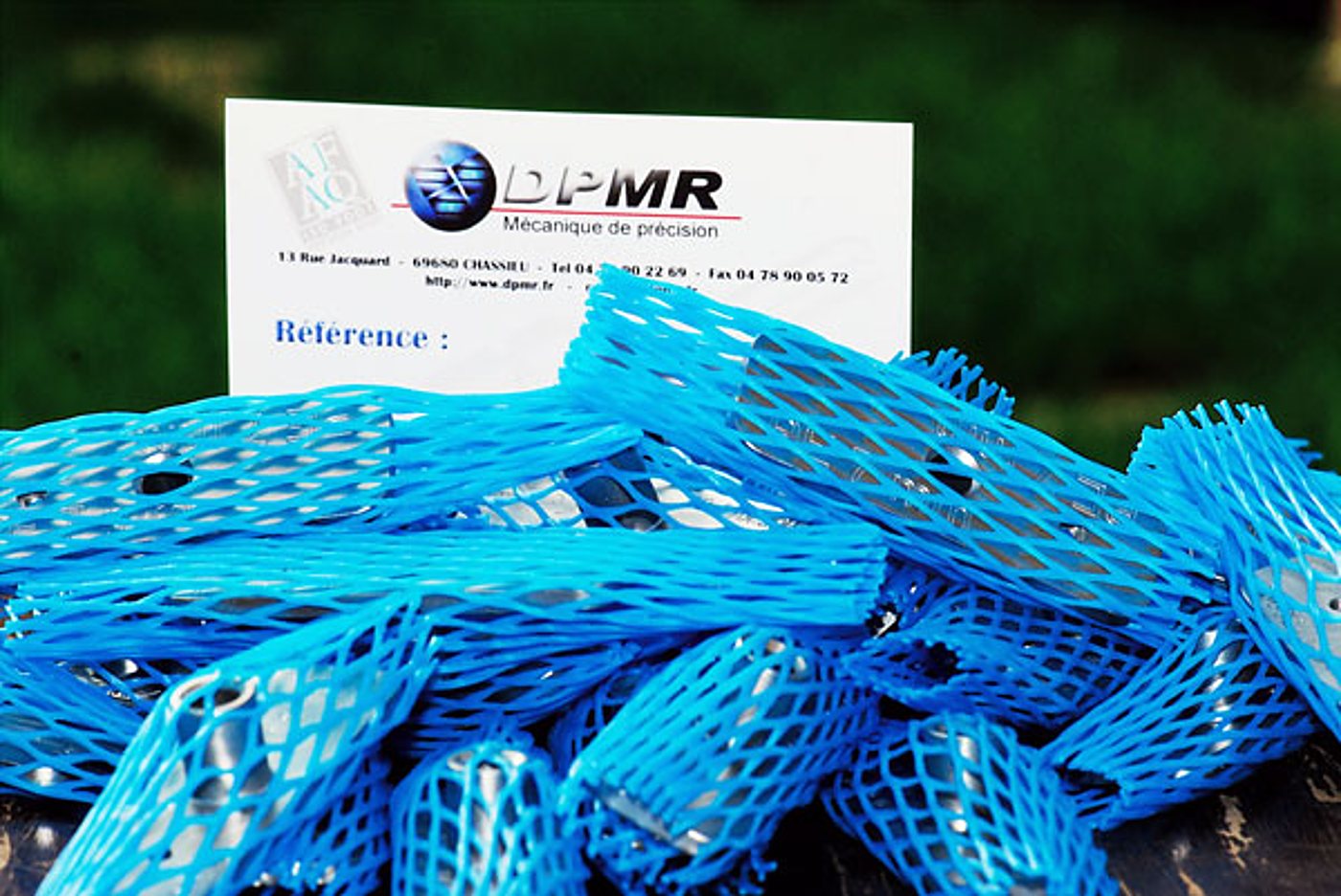 Des pièces mécaniques cylindriques protégées par des filets bleus sont posées devant une carte de visite de l'entreprise DPMR, spécialiste de la mécanique de précision.